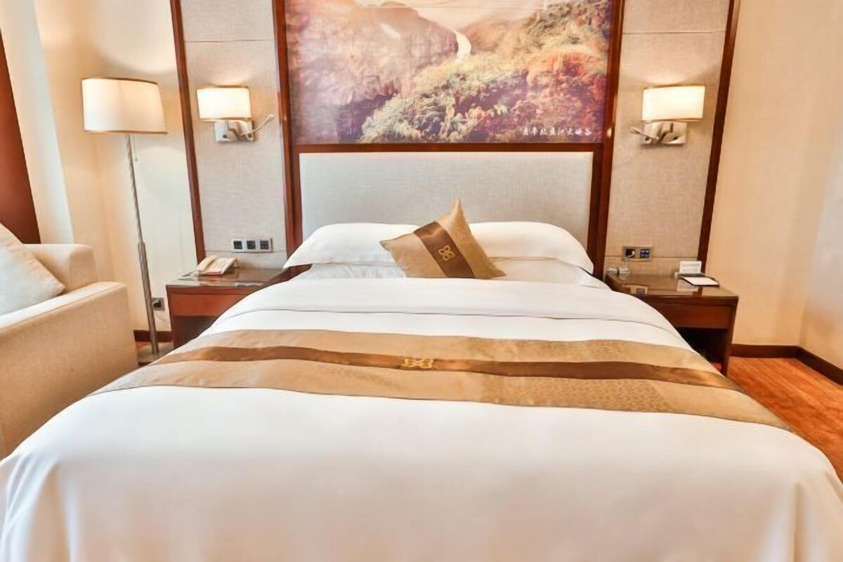 A hotel room with a large bed, two bedside tables, a nightstand, a chair, and a wall-mounted lamp.