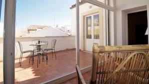 Een balkon met een tafel en stoelen, een rieten stoel en uitzicht op omliggende gebouwen.