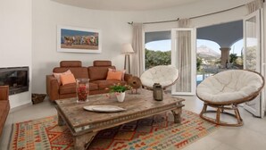 Un salon avec un canapé marron, une table basse et deux fauteuils blancs. Un tableau orne le mur, et on peut admirer, à travers les portes vitrées, une vue sur une montagne et une piscine.