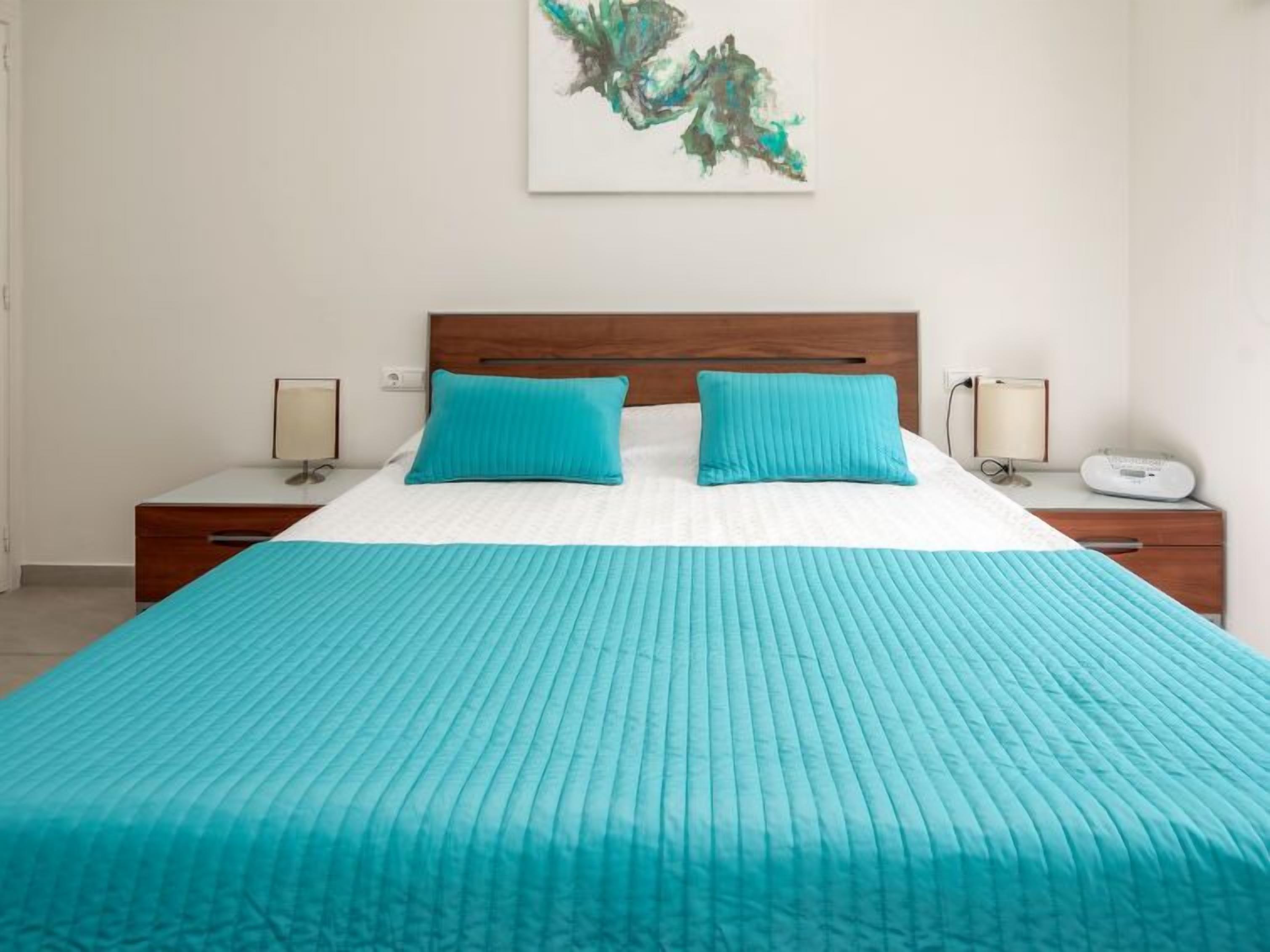 Una cama bien hecha con colcha y almohadas color turquesa, cabecera de madera, mesitas de noche con lámparas y una obra de arte abstracta en la pared.