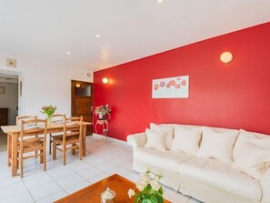 A living room with a red accent wall, a white sofa, wooden dining table with chairs, and a small side table with flowers.