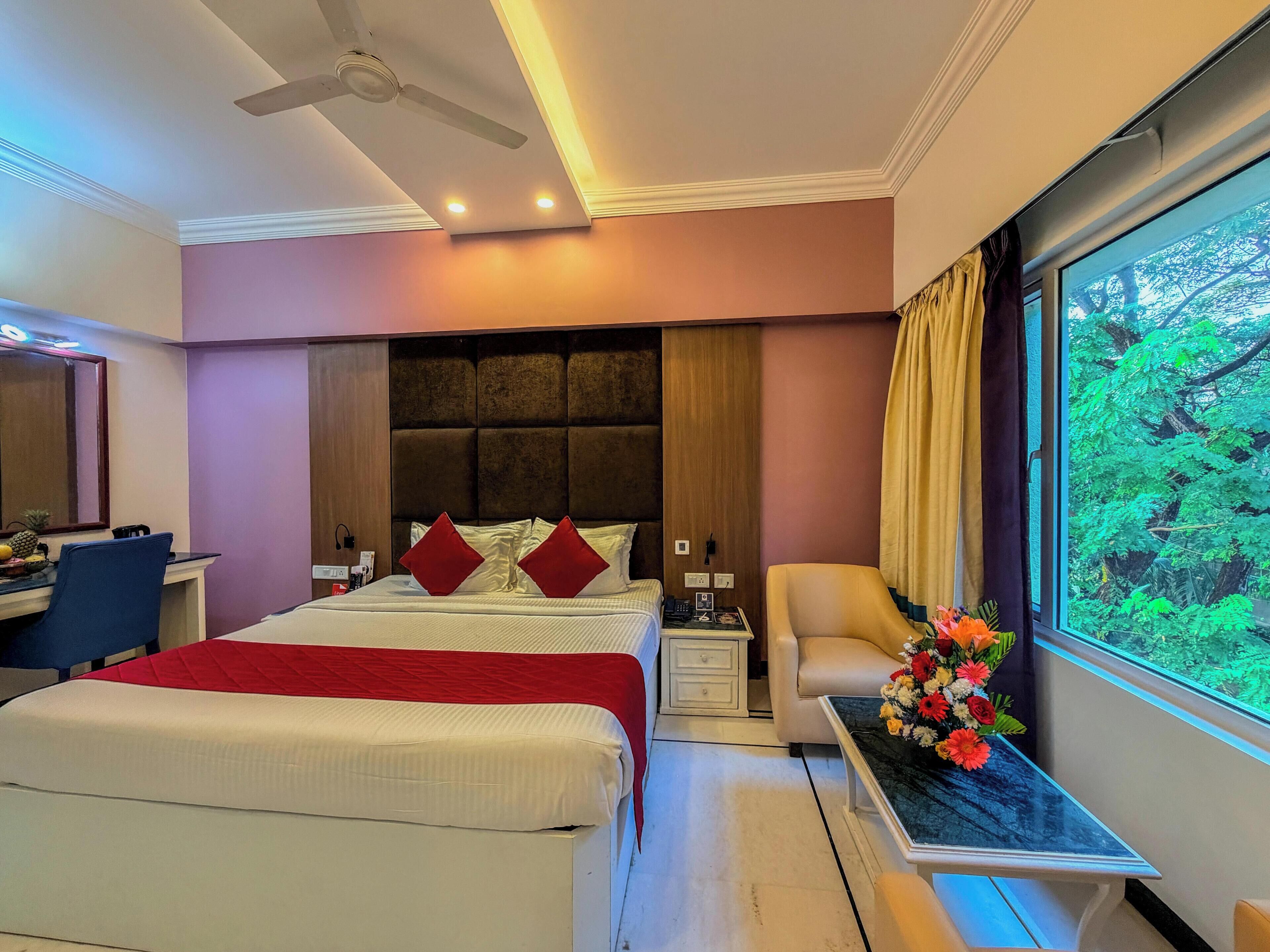A hotel room with a bed, bedside table, chair, desk, and a large window with a view of greenery.