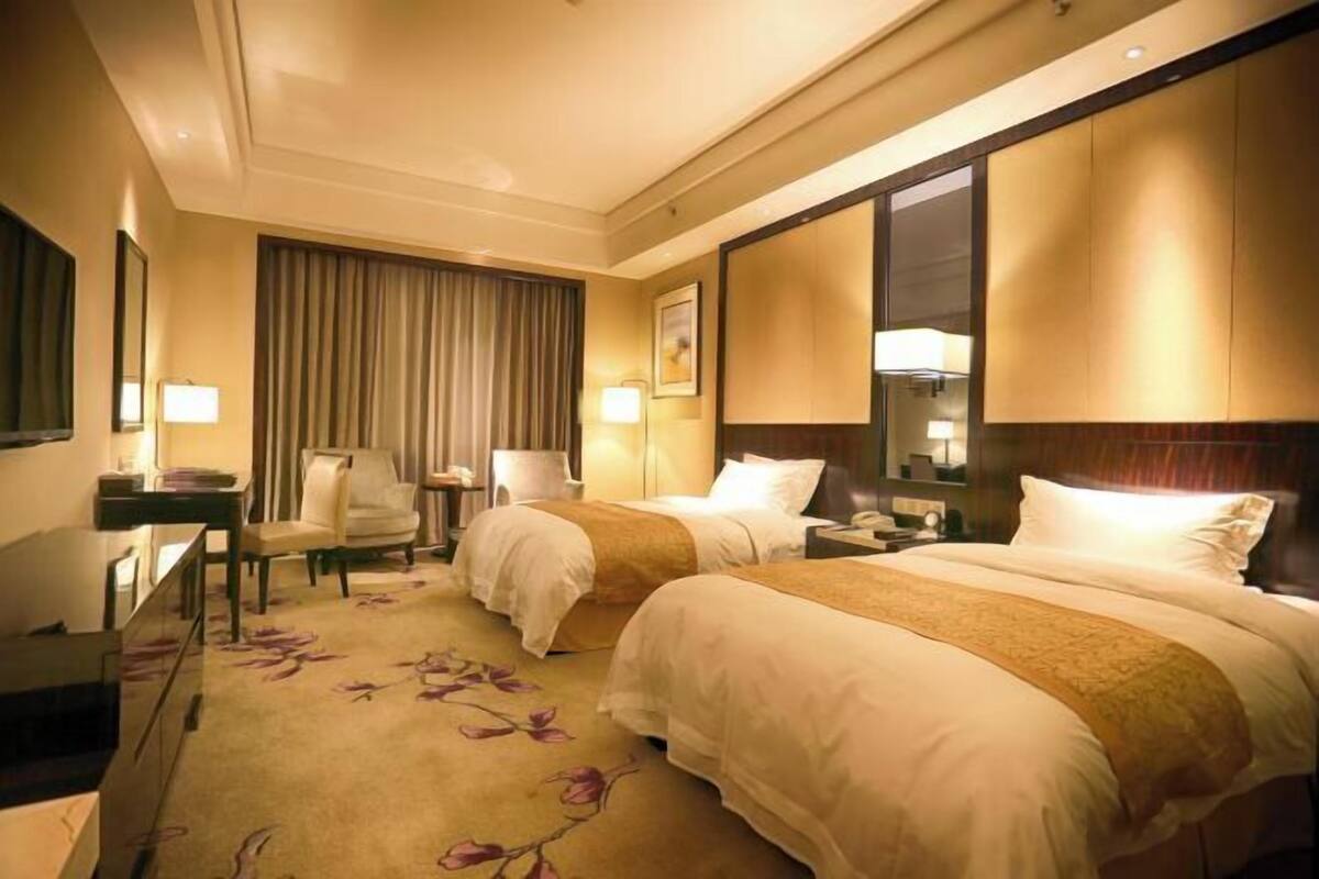 Une chambre d’hôtel avec deux lits, un bureau, une chaise, une grande fenêtre avec des rideaux et une moquette à motifs floraux.