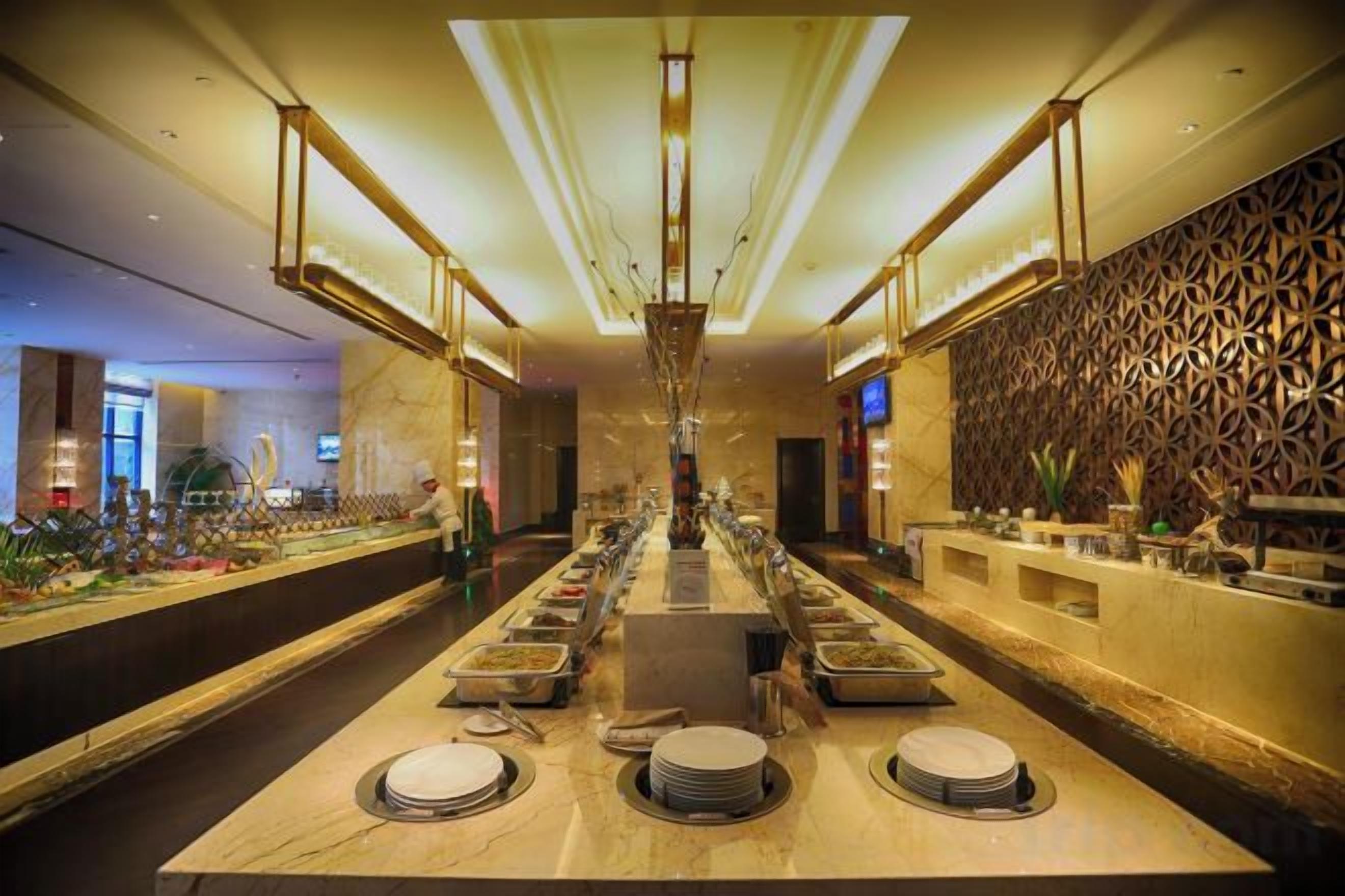 A long dining table with multiple plates and cutlery, surrounded by a modern interior with a patterned wall and overhead lighting.
