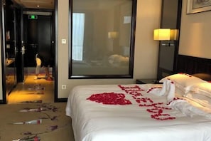 Una habitación de hotel con una cama cubierta de pétalos de rosa, un espejo grande y una mesita de noche con una lámpara.