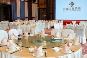 Un salón de banquetes con mesas redondas dispuestas para un evento formal, con una gran pantalla al fondo mostrando la marca del hotel.