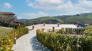 A paved pathway leading to a terrace with seating, surrounded by greenery and overlooking rolling hills.
