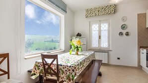 A dining area with a table and chairs, a window with a view of greenery, a radiator, and a kitchen area with cabinets and a sink.