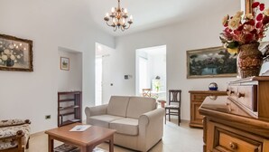 A living room with a sofa, coffee table, and wooden furniture. There is a chandelier, a vase with flowers, and a painting on the wall.