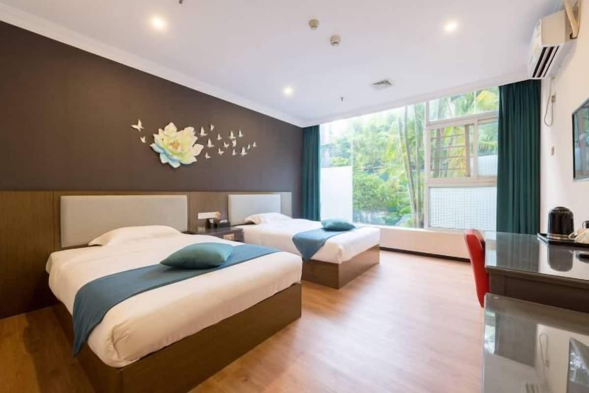 A hotel room with two beds, a desk, and a window with a view of greenery.