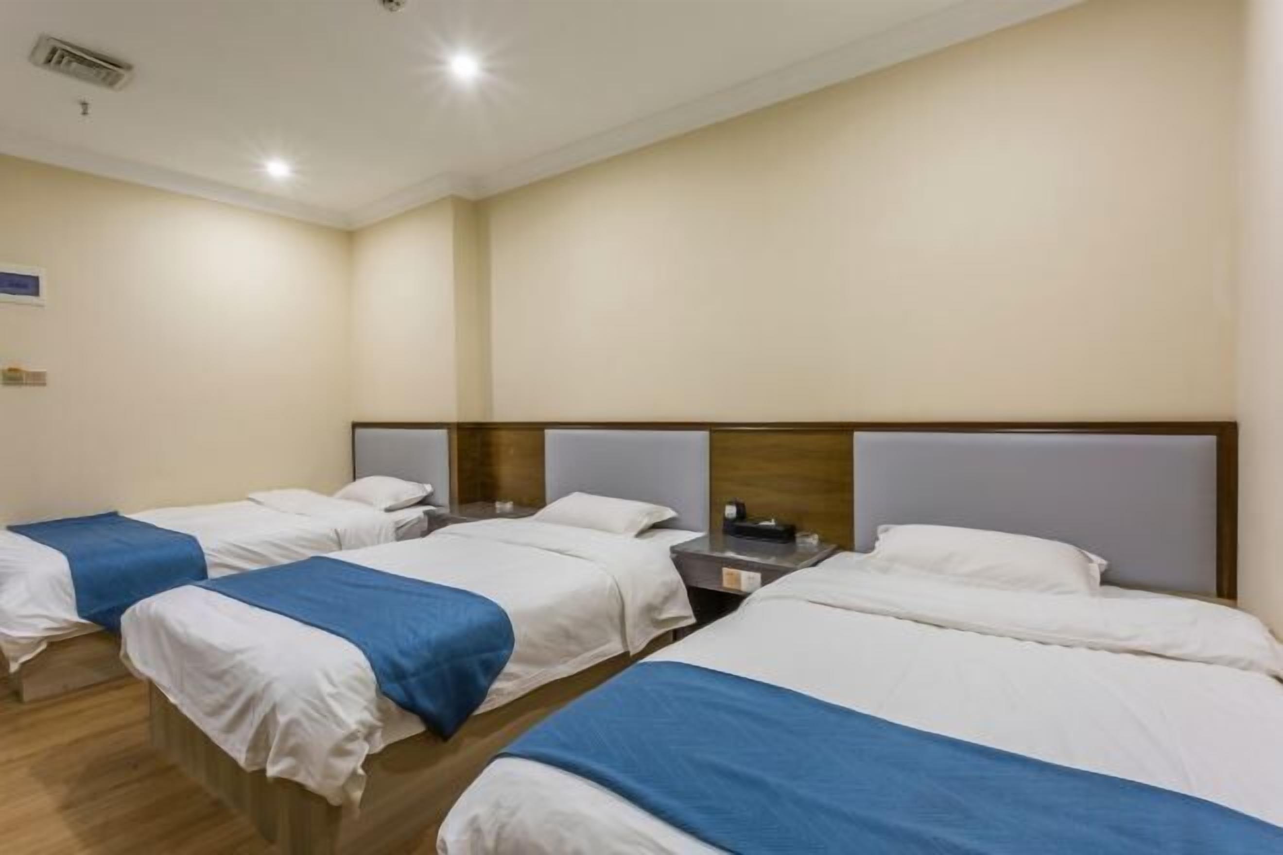 A hotel room with four single beds, each with a wooden headboard and white bedding. There is a small table between the beds with a lamp and a phone.