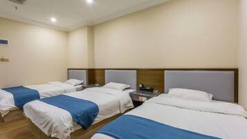 A hotel room with four single beds, each with a wooden headboard and white bedding. There is a small table between the beds with a lamp and a phone.