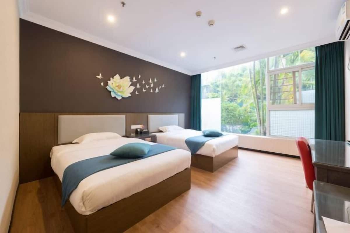 A hotel room with two beds, a desk, a chair, and a view of greenery outside.