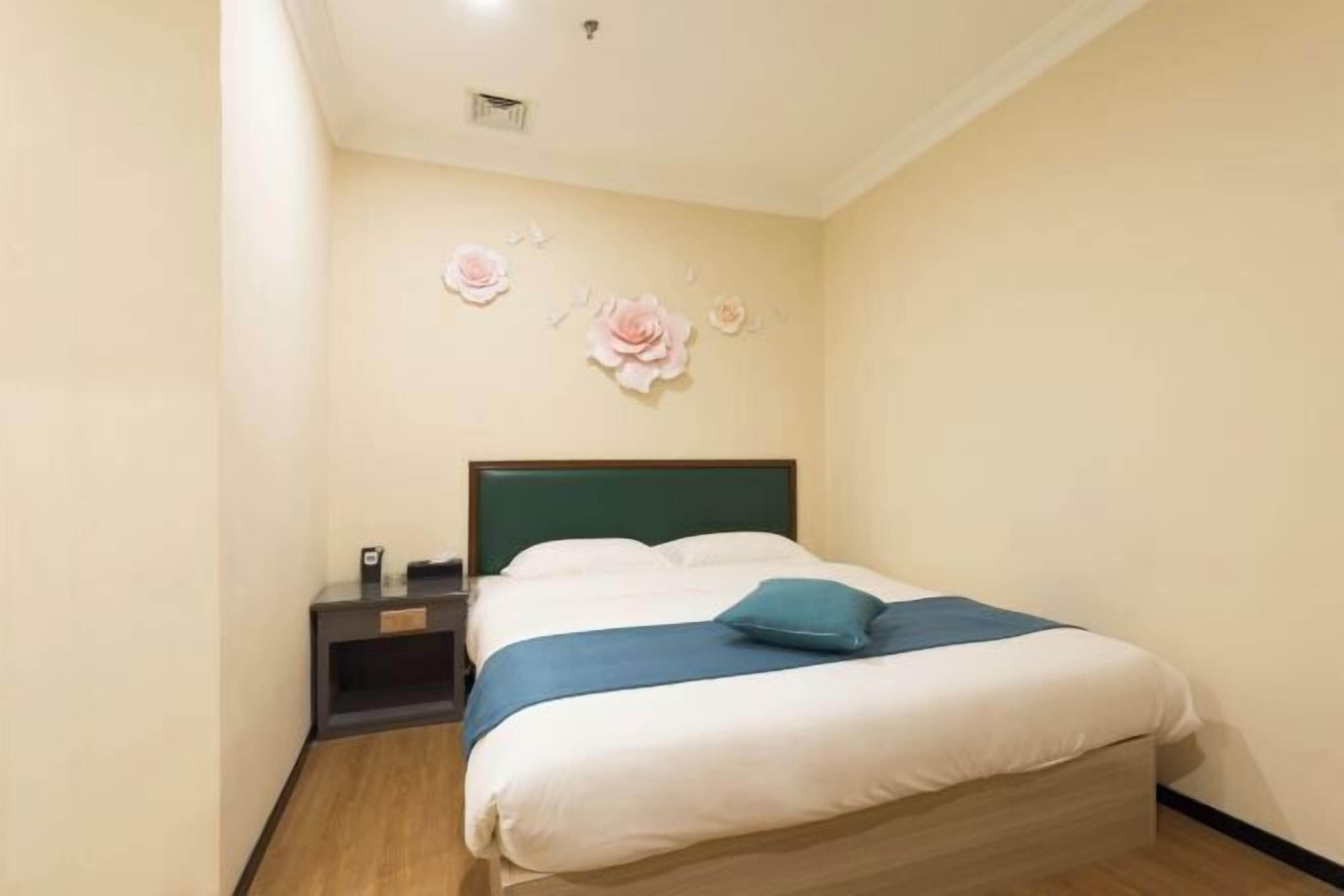 Una cama bien hecha con un almohadón azul y una manta verde azulada, una mesita de noche con una lámpara y decoración en la pared.