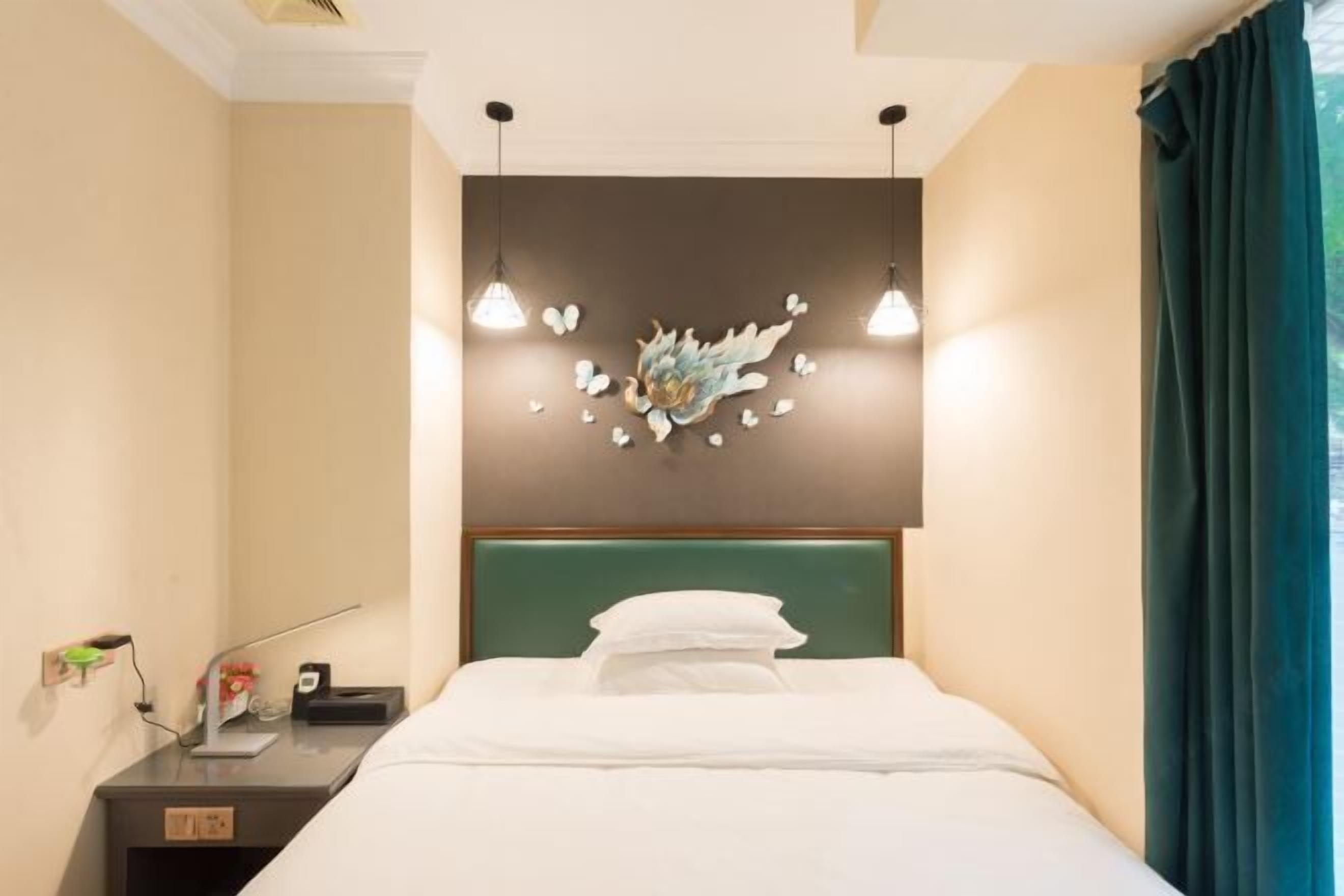 A hotel room with a bed, a nightstand, a wall-mounted light fixture, and a window with curtains.