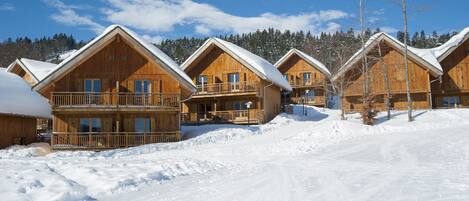 A row of wooden cabins with balconies, surrounded by snow-covered ground and trees.
