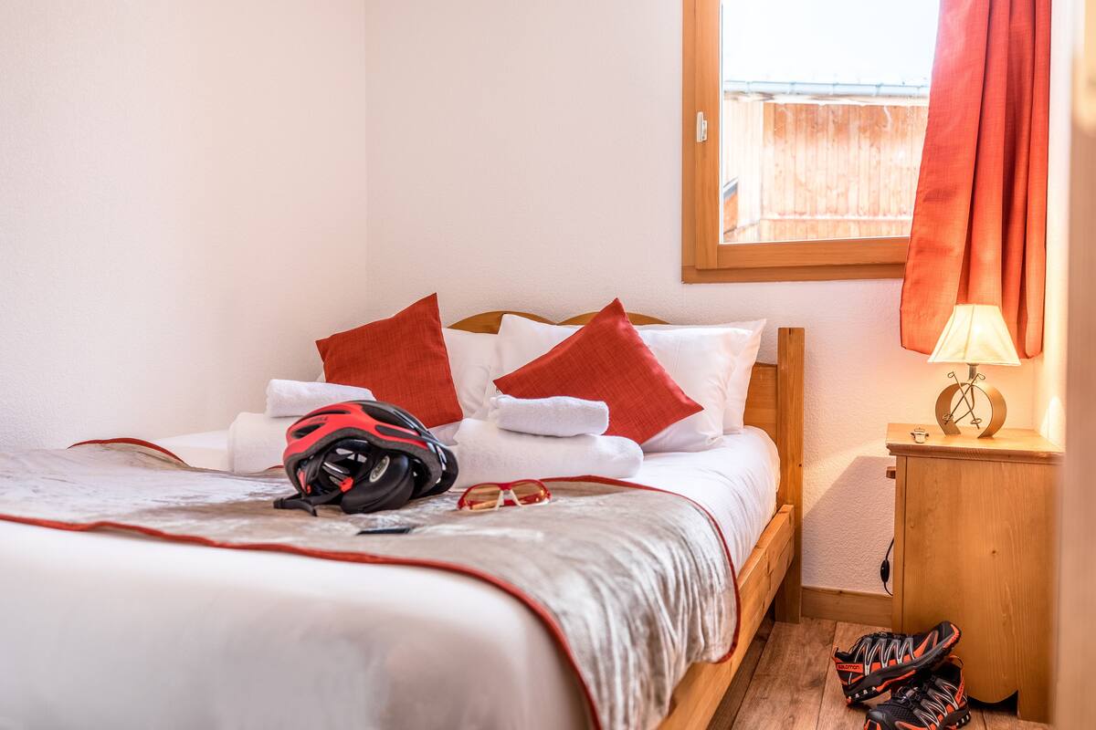 Ein ordentlich bezogenes Bett mit einem roten und weißen Kissenpaar, einem roten Rucksack und einer Sonnenbrille darauf. Ein Holz-Nachttisch mit einer Lampe und ein Paar Schuhe stehen auf dem Boden neben dem Bett.