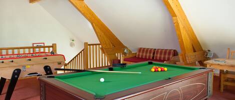 A pool table with balls, a foosball table, and a sofa in a room with wooden beams.