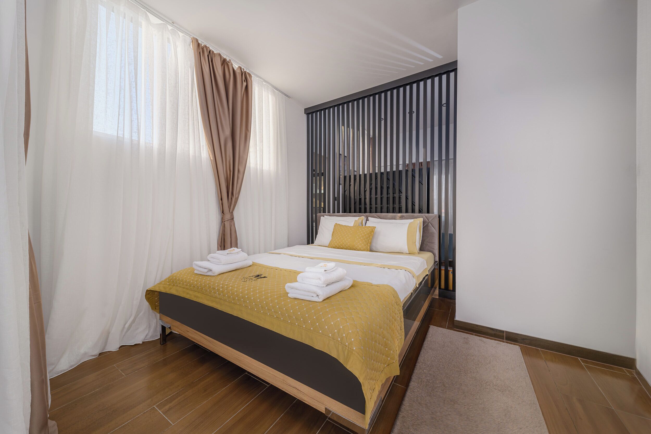 A hotel room with a bed, curtains, a headboard, and a wooden floor.