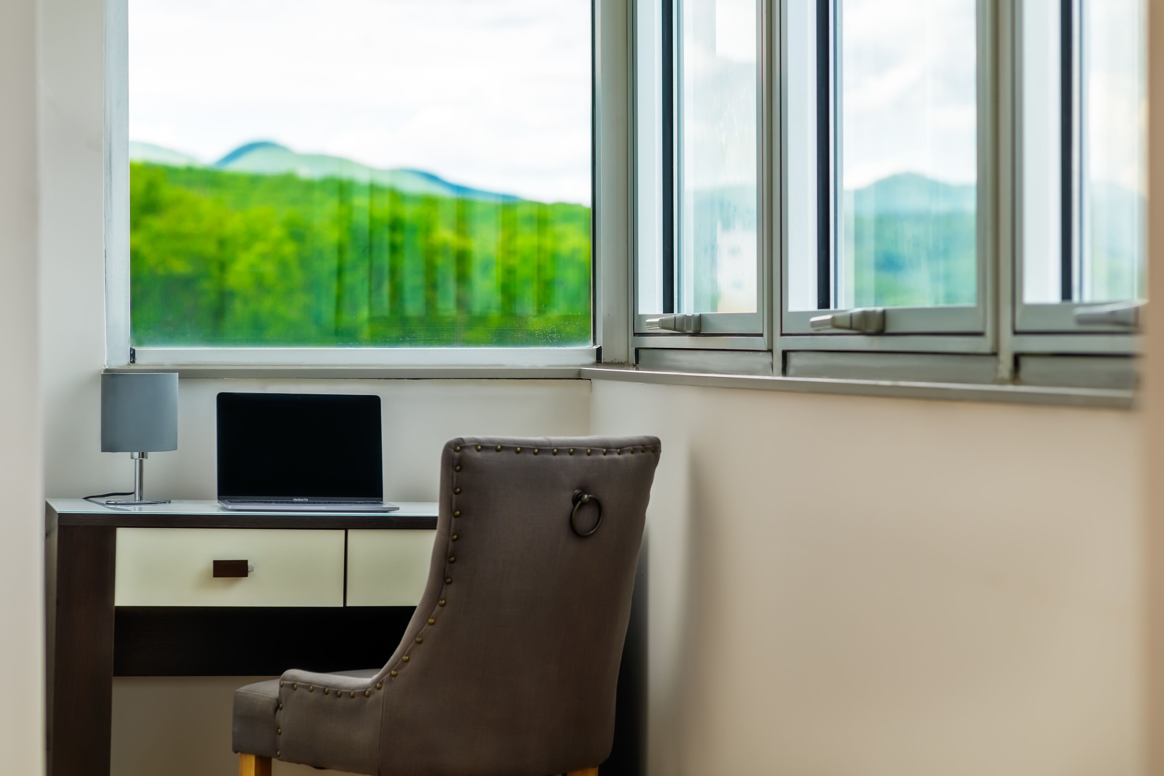 A room with a desk, chair, and a window with a view of greenery and mountains.