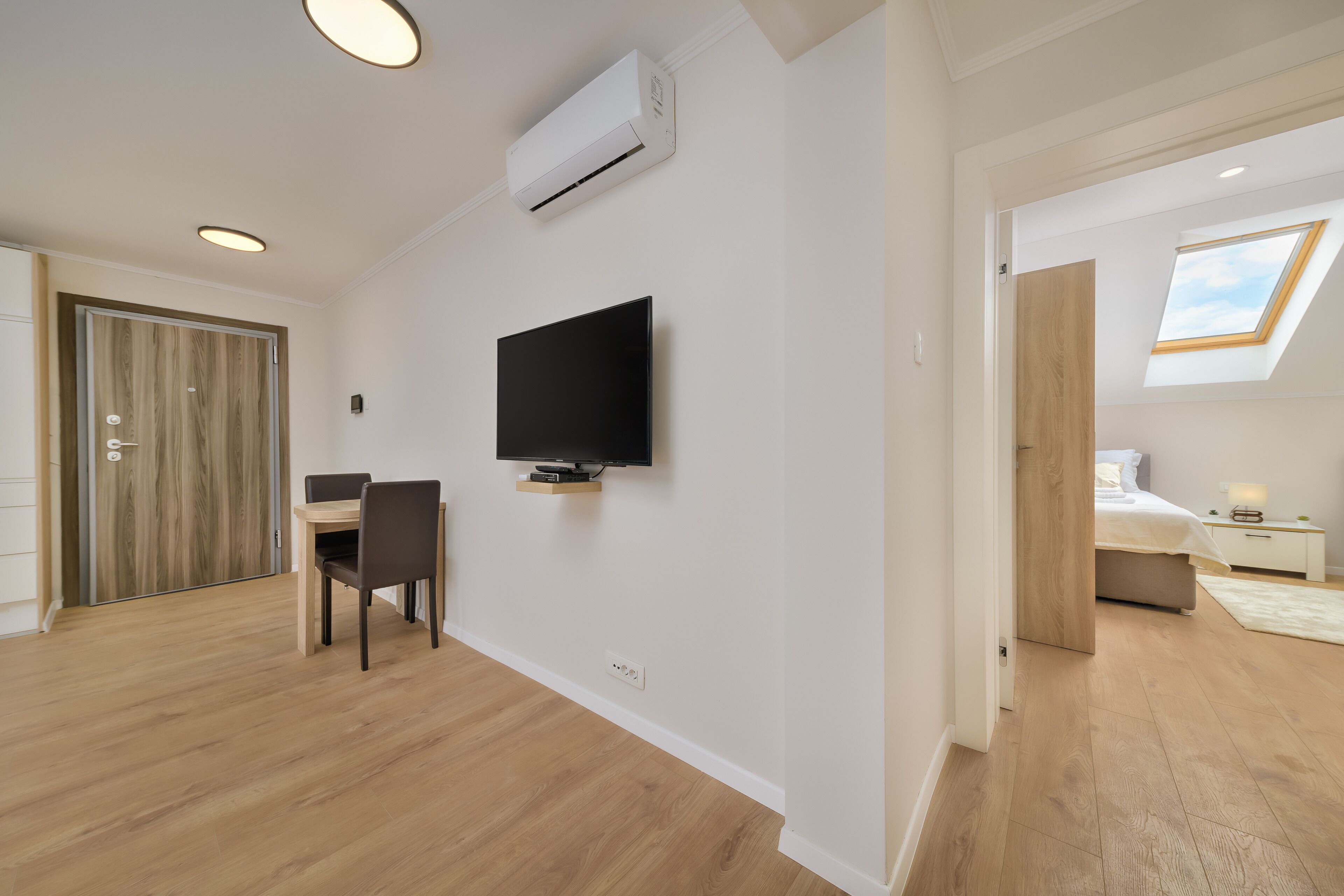 A modern room with a flat-screen TV mounted on the wall, a wooden dining table with chairs, and a skylight in the adjacent bedroom.