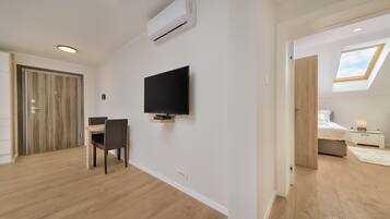 A modern room with a flat-screen TV mounted on the wall, a wooden dining table with chairs, and a skylight in the adjacent bedroom.