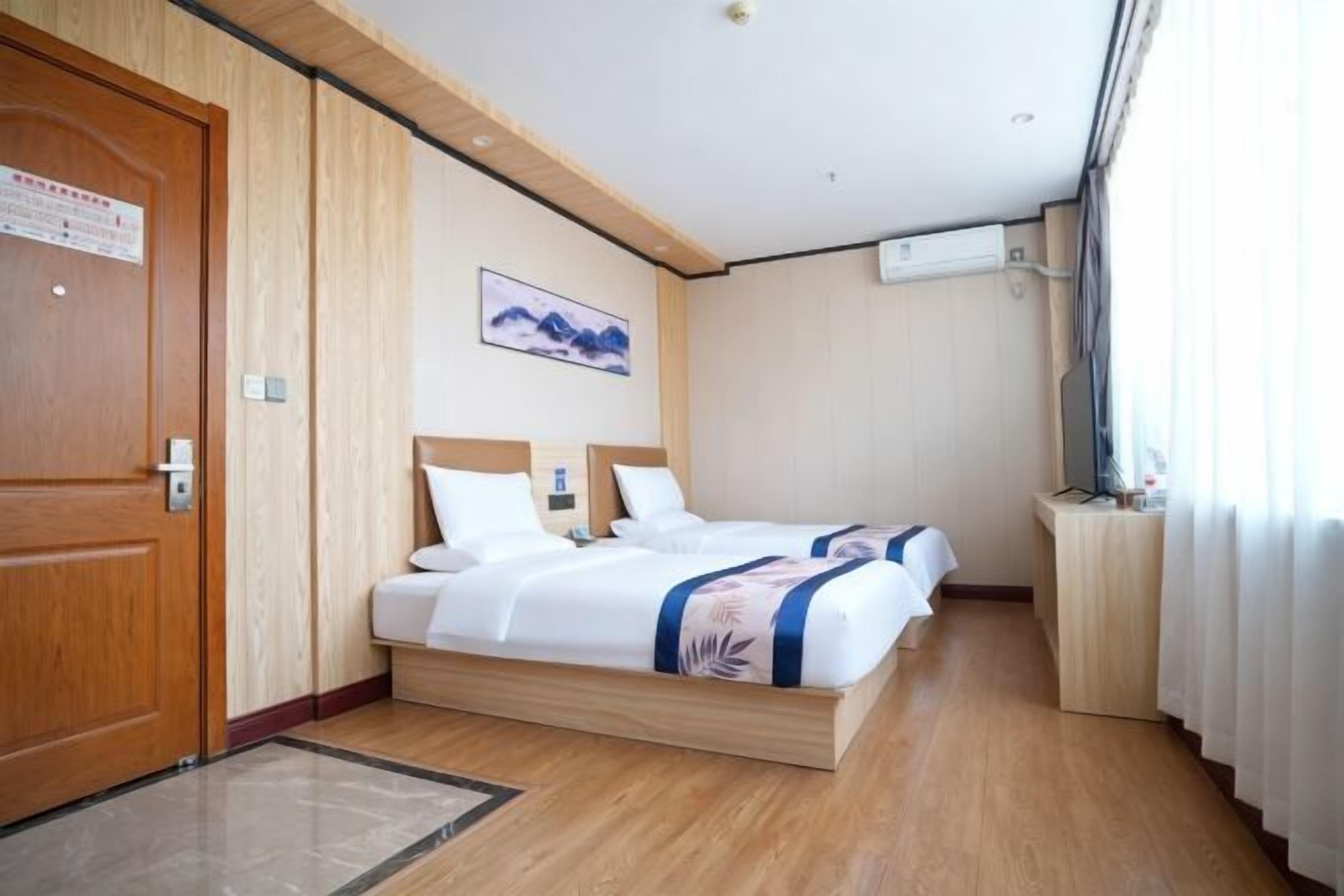 Quarto de hotel com porta de madeira, cama com roupa de cama branca, quadro na parede e pequena escrivaninha com luminária.