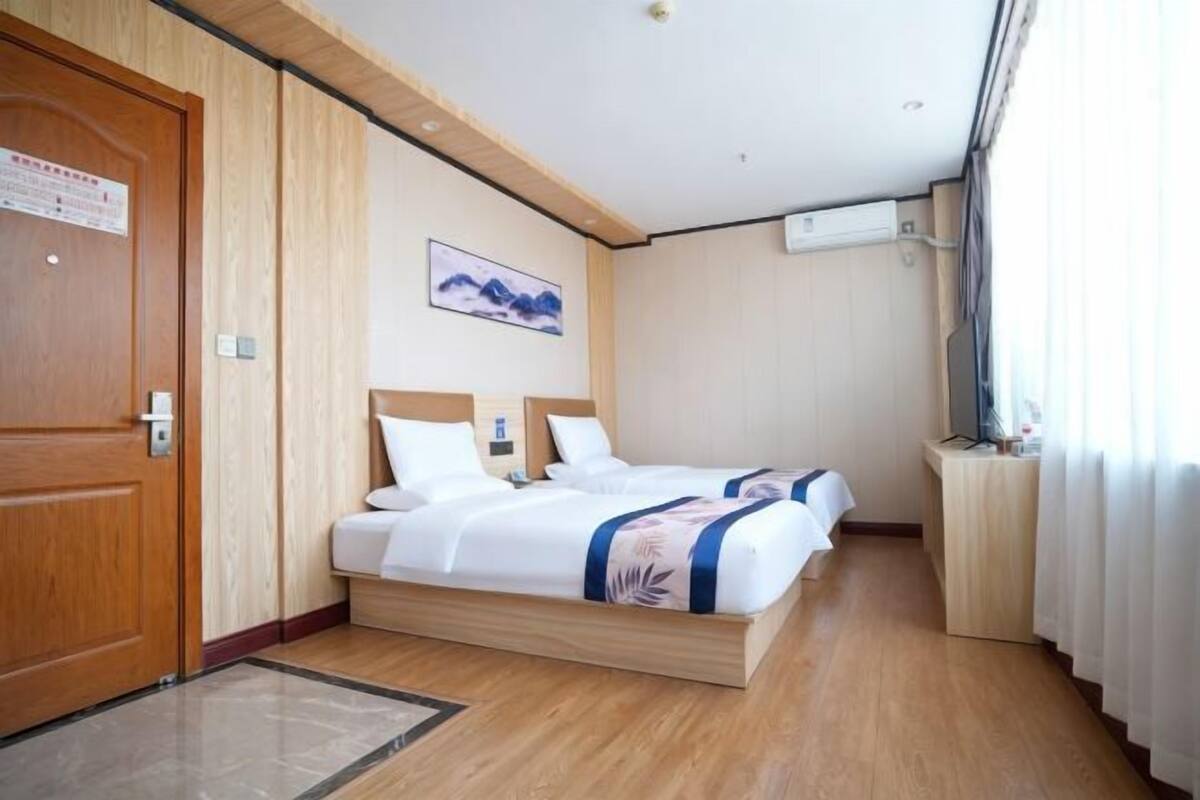 A hotel room with a wooden door, a bed with white bedding, a painting on the wall, and a small desk with a lamp.