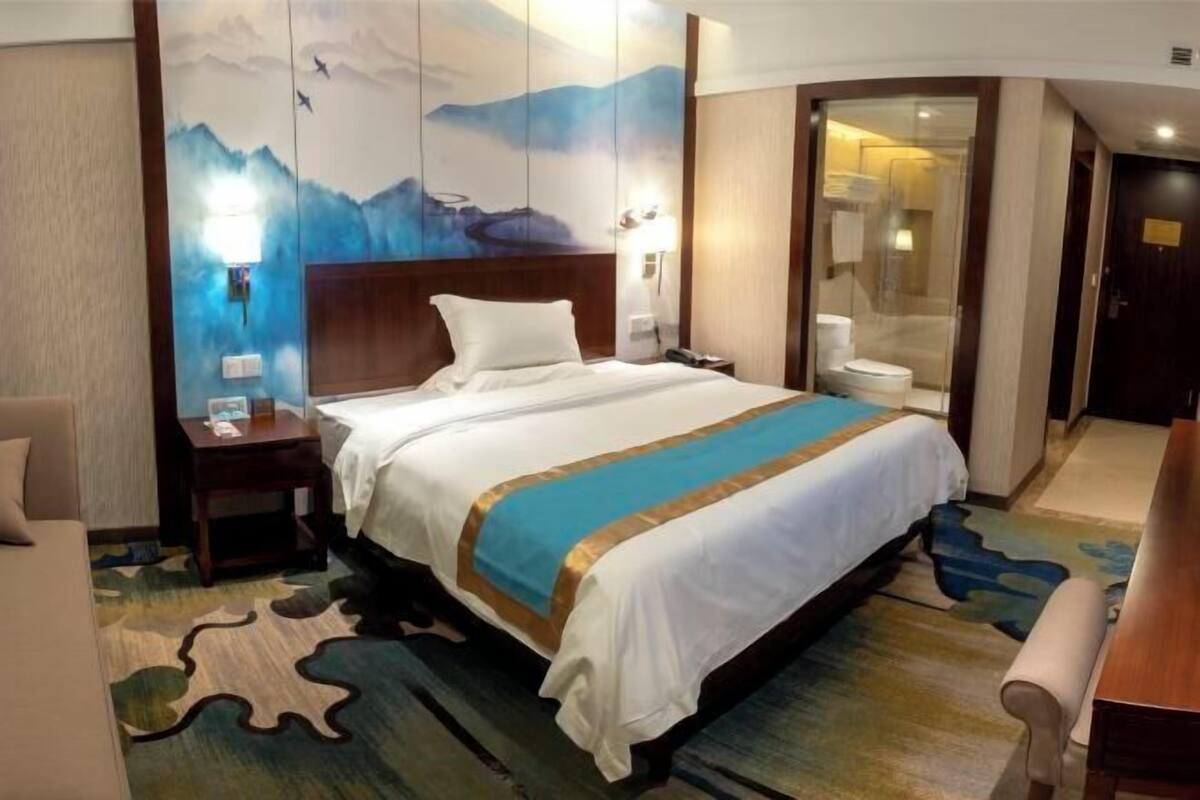 A hotel room with a large bed, a nightstand, a desk, a chair, a bathroom with a glass door, and a wall with a mountain painting.