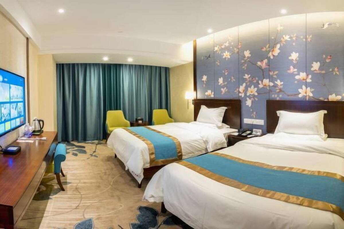 A hotel room with two beds, a desk with a flat-screen TV, and a large window with curtains.