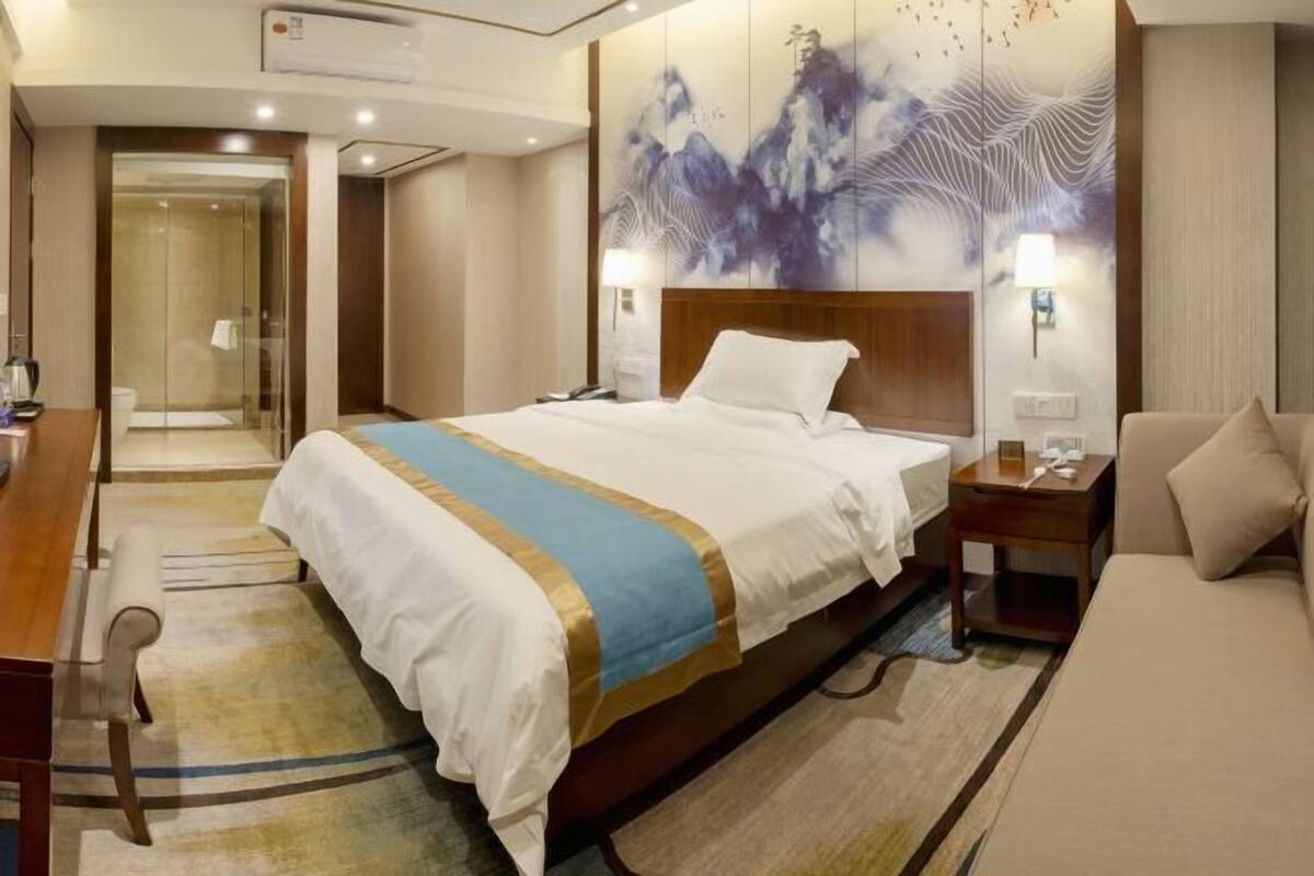A hotel room with a large bed, a bedside table, a nightstand, a sofa, and a bathroom with a glass door.