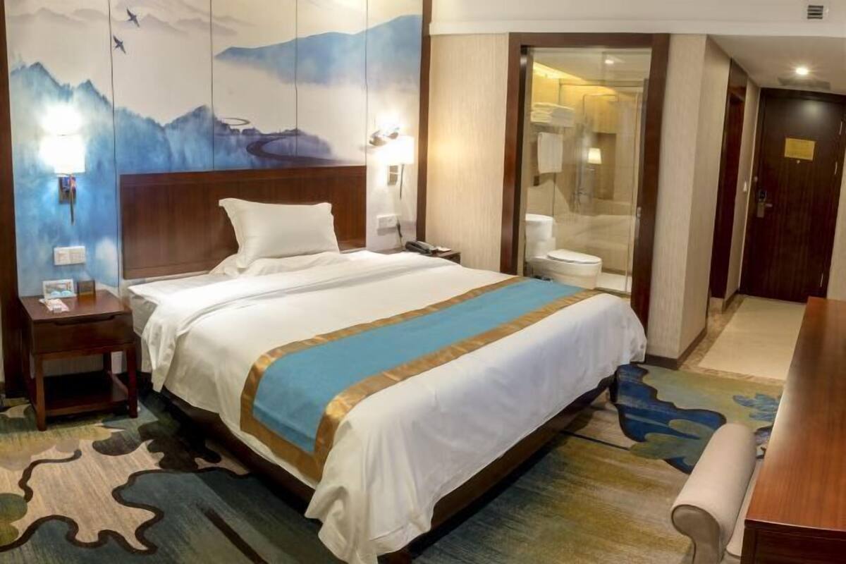 A hotel room with a large bed, a wooden headboard, a nightstand, a bathroom with a glass partition, and a view of the mountains.