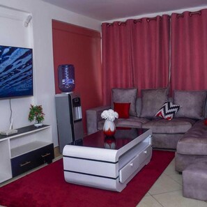 A living room with a grey sofa, a coffee table, a flat-screen TV, a water dispenser, and a red curtain. - Stay.Plus Kasarani Budget Apartment (Nairobi)