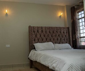 A bedroom with a tufted headboard, two wall-mounted lights, a bed with a white bedspread, and a window with curtains. - Stay.Plus Kasarani Budget Apartment (Nairobi)