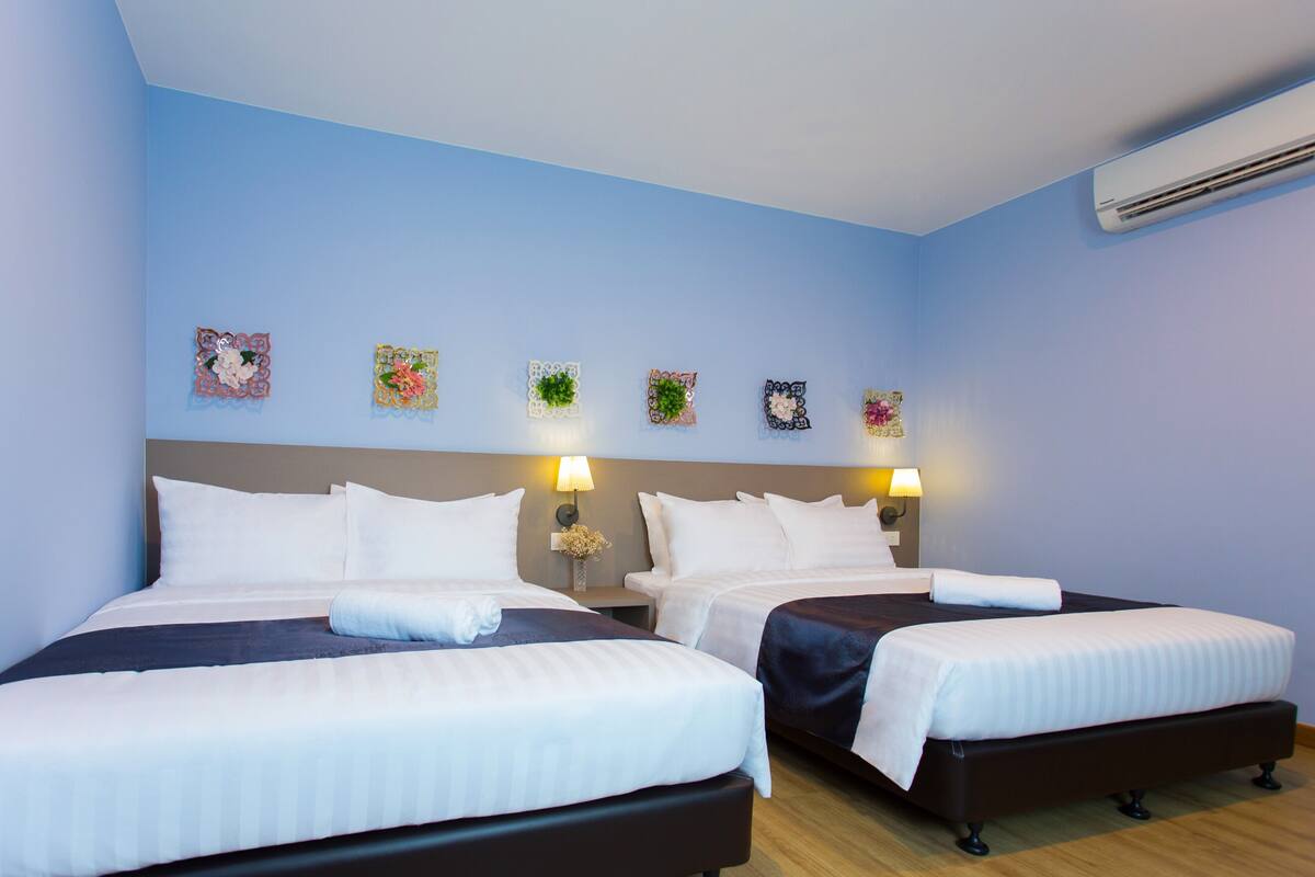 A hotel room with two beds, a nightstand with a lamp, decorative wall art, and an air conditioning unit.