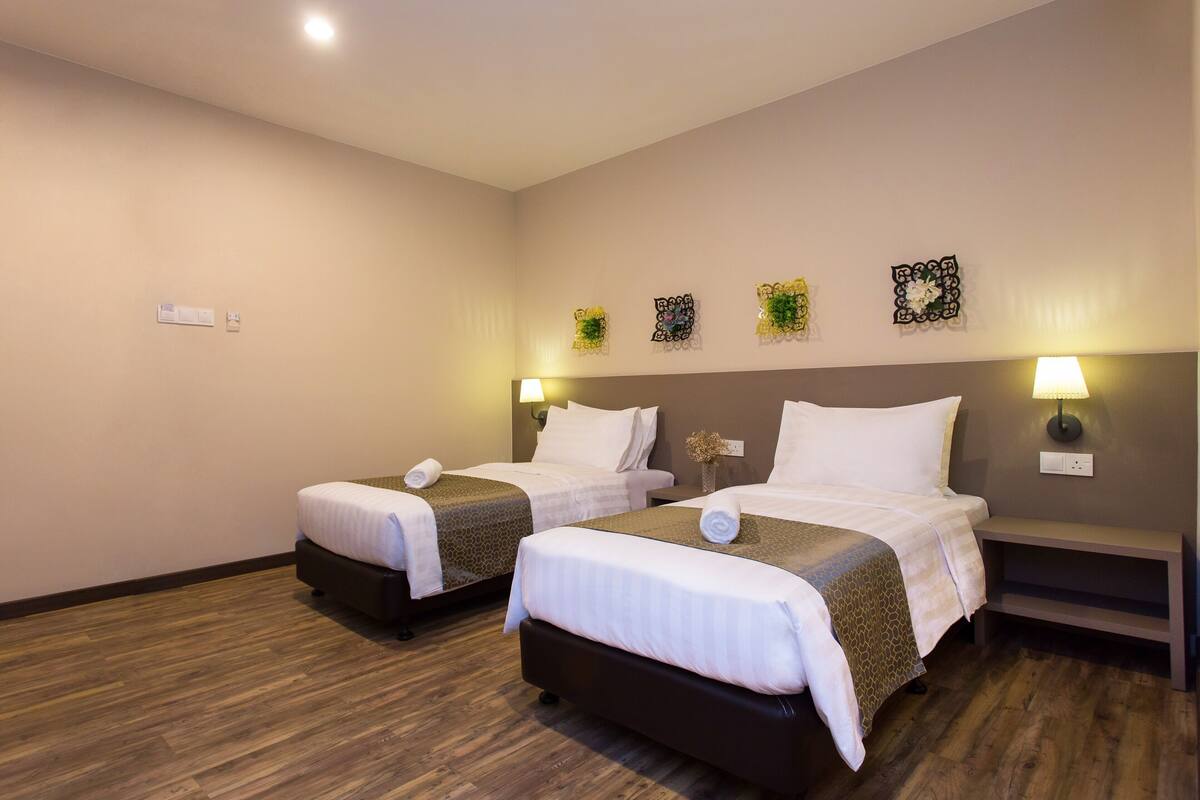 A hotel room with two beds, wooden flooring, and wall-mounted lamps.
