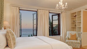 A bedroom with a large bed, two open windows with curtains, a chair, and a chandelier. - Buccara Noetzie - Pezula Castle (Knysna)