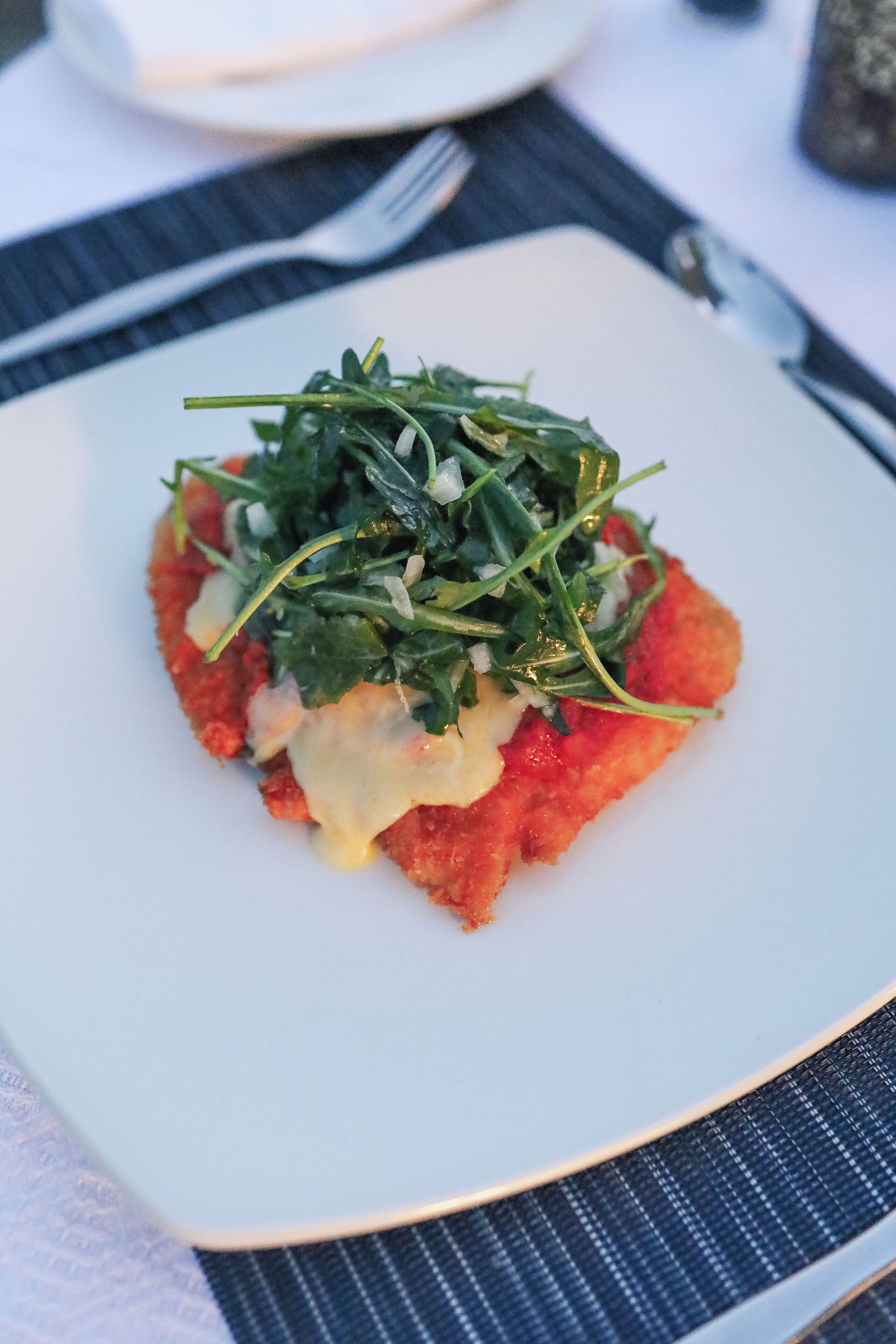 A square plate with a red food item topped with green leaves and cheese.