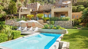 A pool with lounge chairs and umbrellas, surrounded by a stone building with balconies and a lush garden. - Buccara Noetzie - Pezula Castle (Knysna)