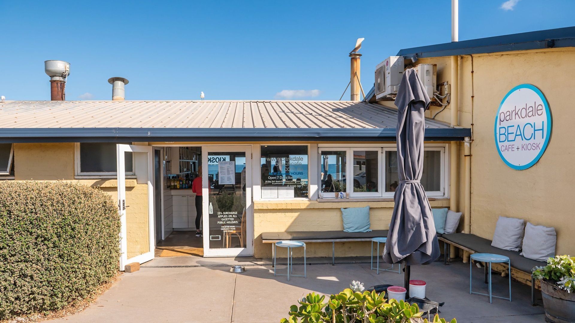 A beachside cafe with outdoor seating, a sign indicating it's a parkdale beach cafe and kiosk, and a clear sky above.