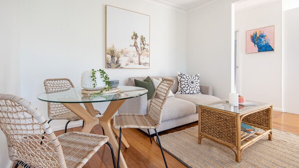 A modern living room with a glass dining table, wicker chairs, a grey sofa, and a small side table.