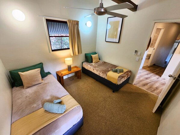 A bedroom with two beds, a ceiling fan, a window with curtains, a bedside table with a lamp, and a door leading to another room.
