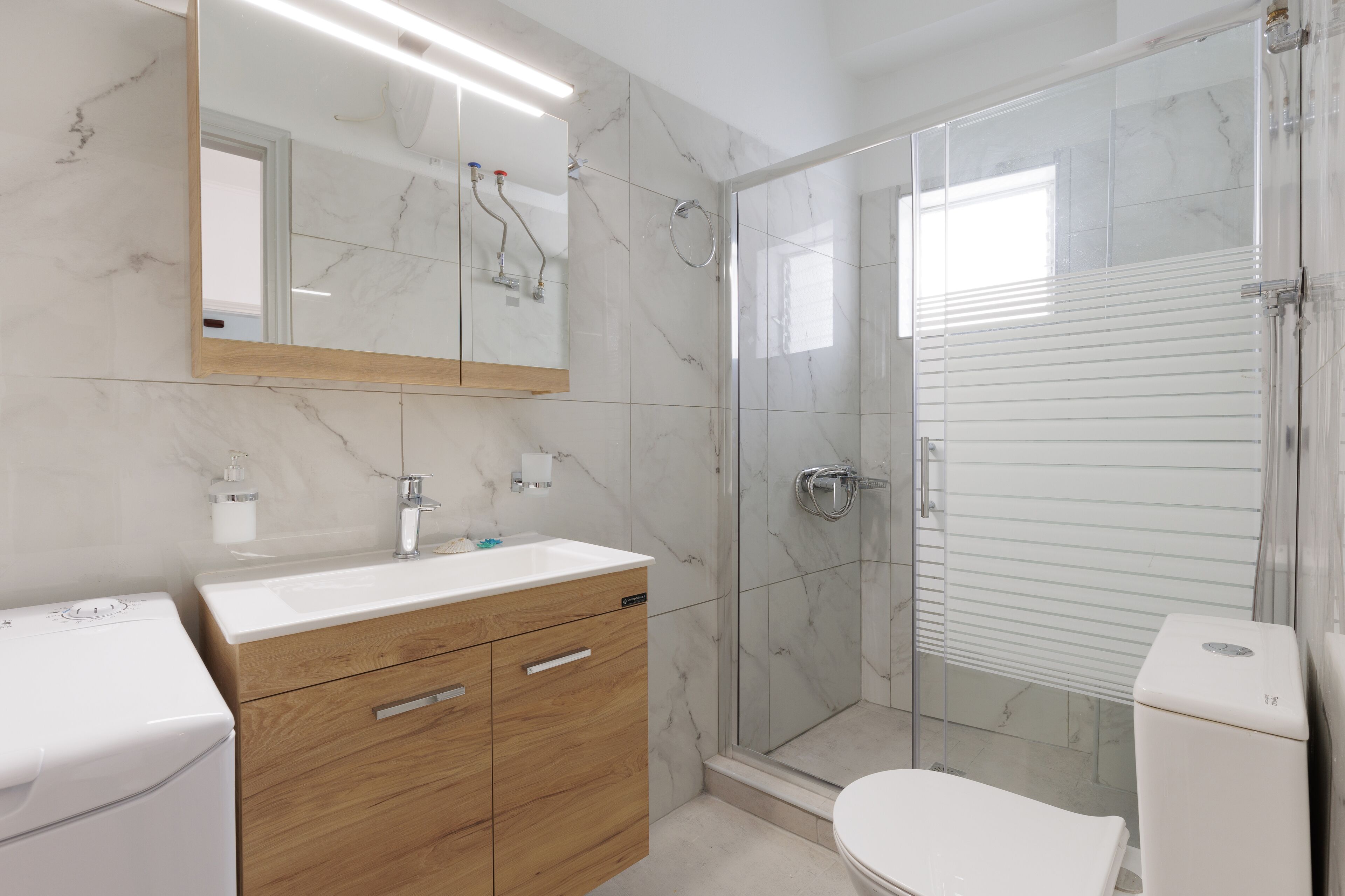 A modern bathroom with a marble backsplash, a wooden vanity, a glass shower enclosure, and a toilet.