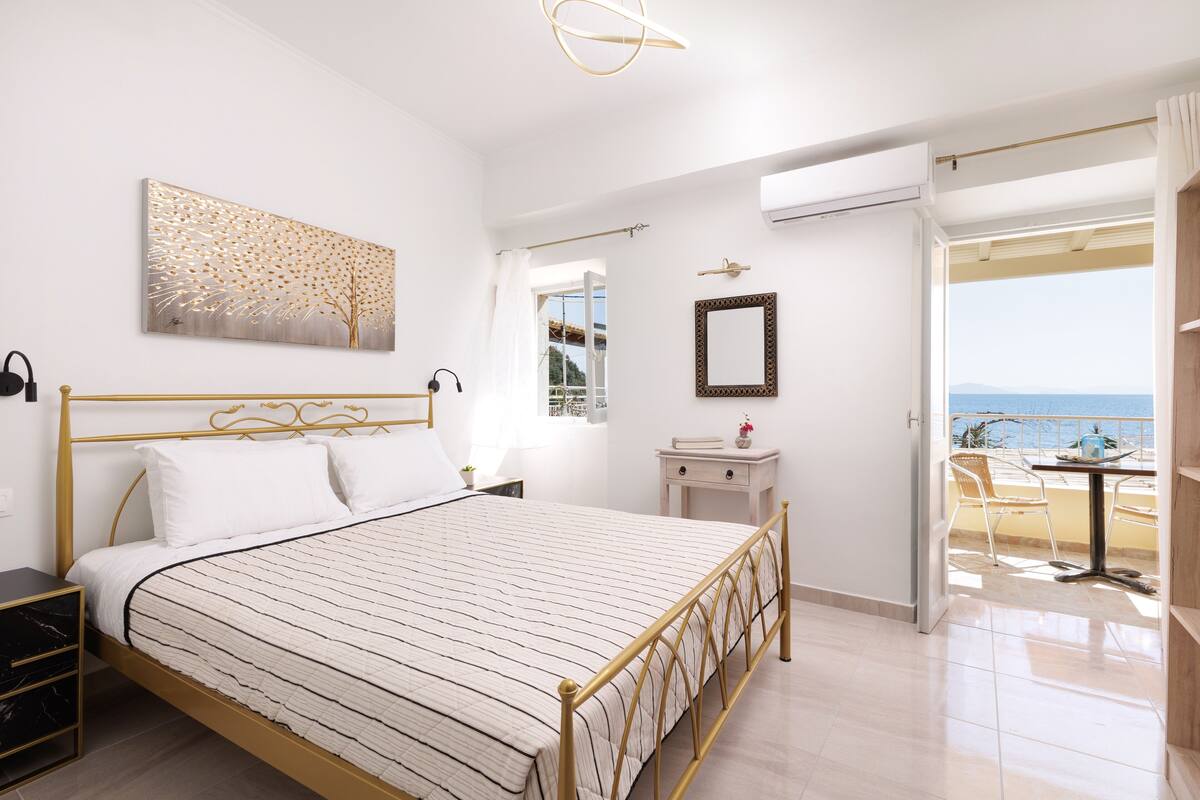 Une chambre à coucher avec un lit, une table de chevet et une vue sur la mer, visible par une porte ouverte.