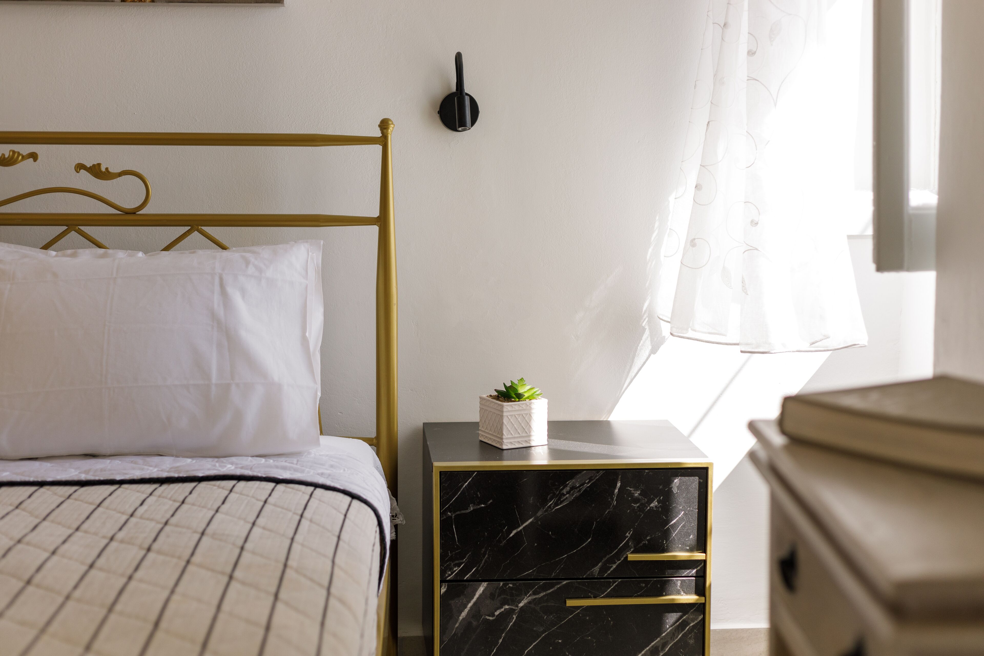 Una cama bien arreglada con cabecera dorada, una mesita de noche con cajones y una lámpara fijada a la pared.