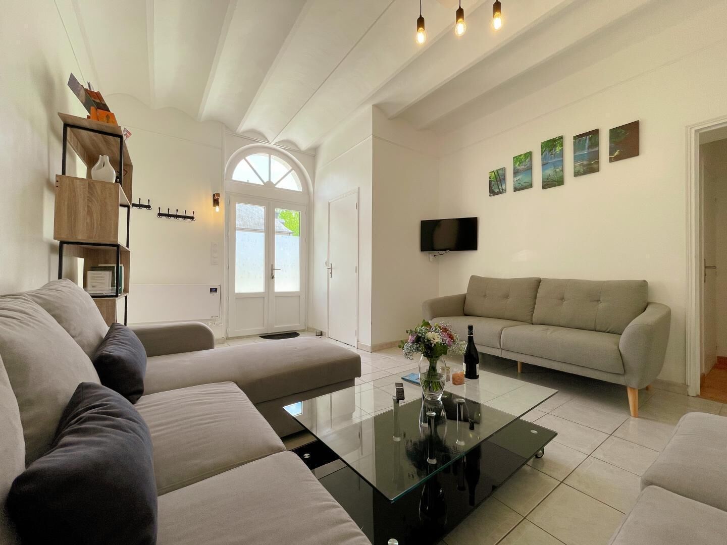 A modern living room with a glass coffee table, beige sofas, a flat-screen TV, and a bookshelf.