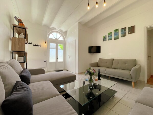 A modern living room with a glass coffee table, beige sofas, a flat-screen TV, and a bookshelf.