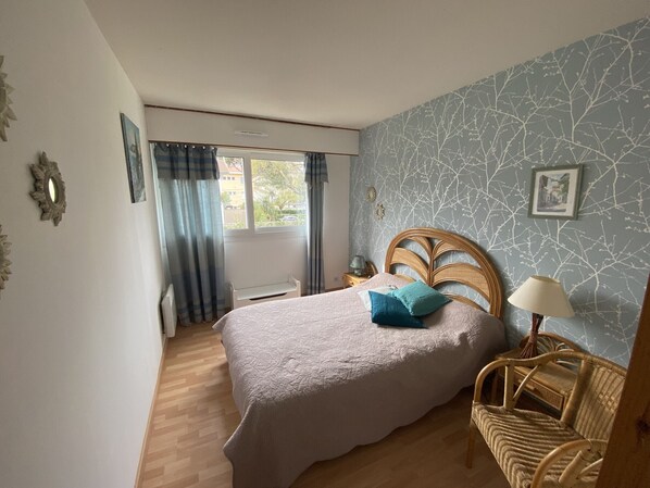 A bedroom with a bed, a wicker chair, a nightstand, a window with curtains, and a wall with a tree branch pattern.