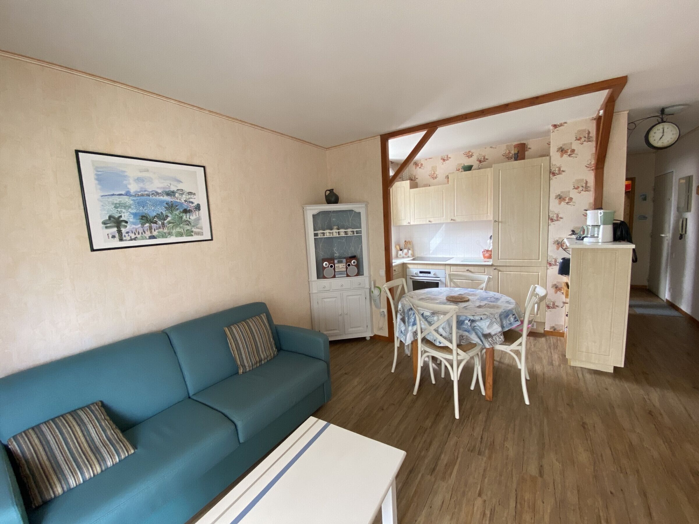 Ein gemütliches Wohnzimmer mit einem blauen Sofa und einem Esstisch mit Stühlen. Ein Küchenbereich ist sichtbar, mit Schränken und einer Uhr.