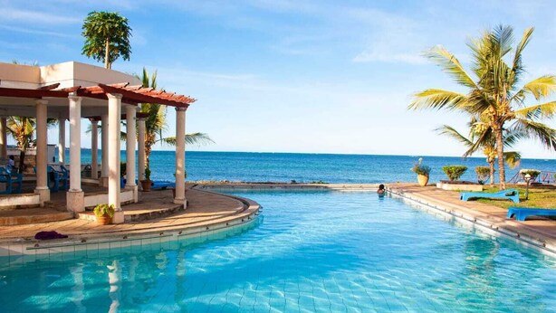 A pool with a view of the ocean, a covered area with chairs, and palm trees. - Stay.Plus Almasi Beachfront Apartments (Mombasa)